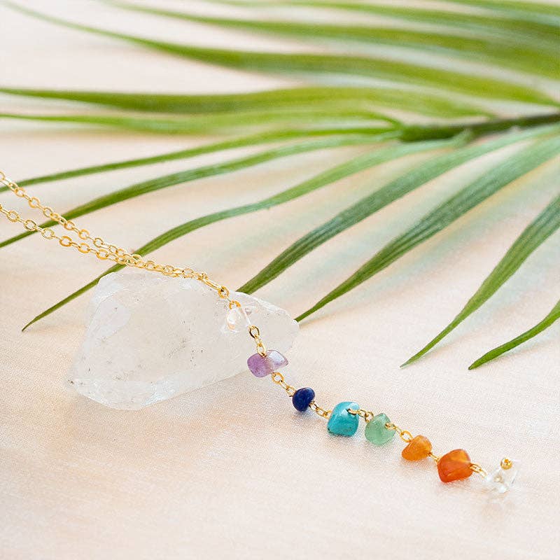 7 Chakras Healing Necklace displayed on a white crystal with palm leaf, featuring natural gemstone beads on a yellow gold chain.