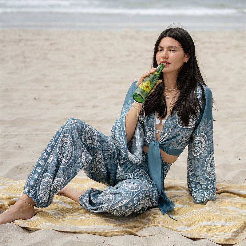 Woman in a blue patterned outfit sitting on a yellow towel at the beach, drinking from a green bottle.