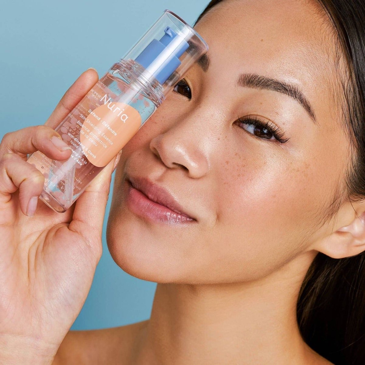 Woman holding Nuria Defend Skin Shielding Essence Mini, a hydrating face essence with niacinamide beads and ginseng for antioxidant skin protection.