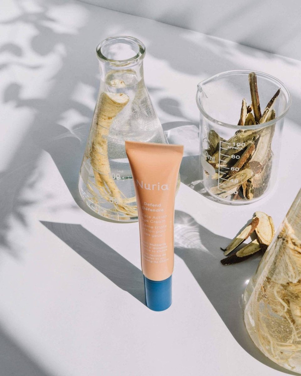 Nuria Eye Cream tube placed next to beakers with natural extracts, highlighting ingredients like Mulberry Root and Ginseng.