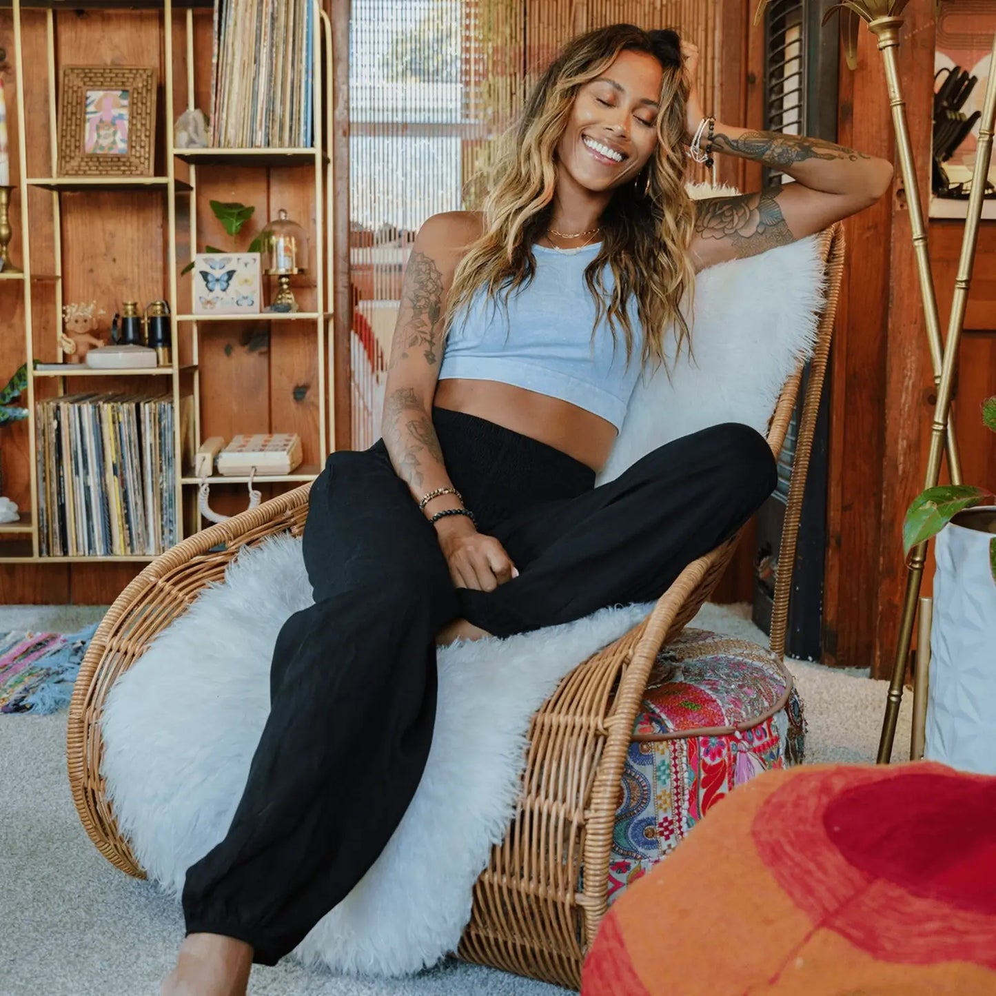 Woman wearing black harem pants sitting on a wicker chair in a cozy room with wooden shelves and plants.