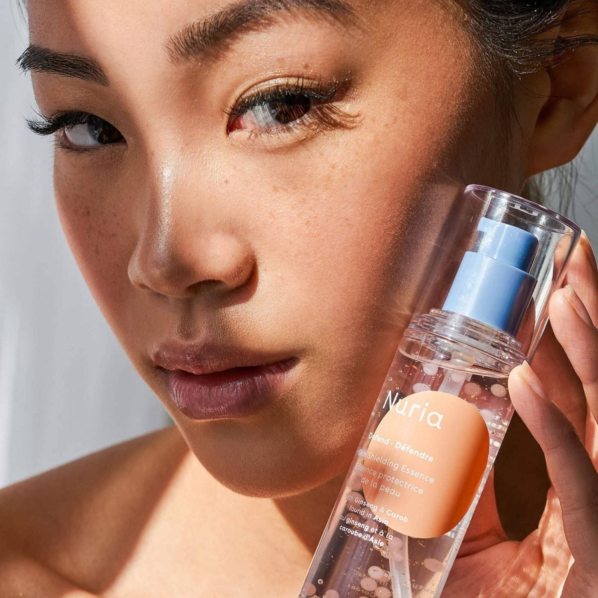Nuria Defend Skin Shielding Essence Mini hydrating face serum with visible niacinamide beads held by model. Clean beauty antioxidant skincare for travel.