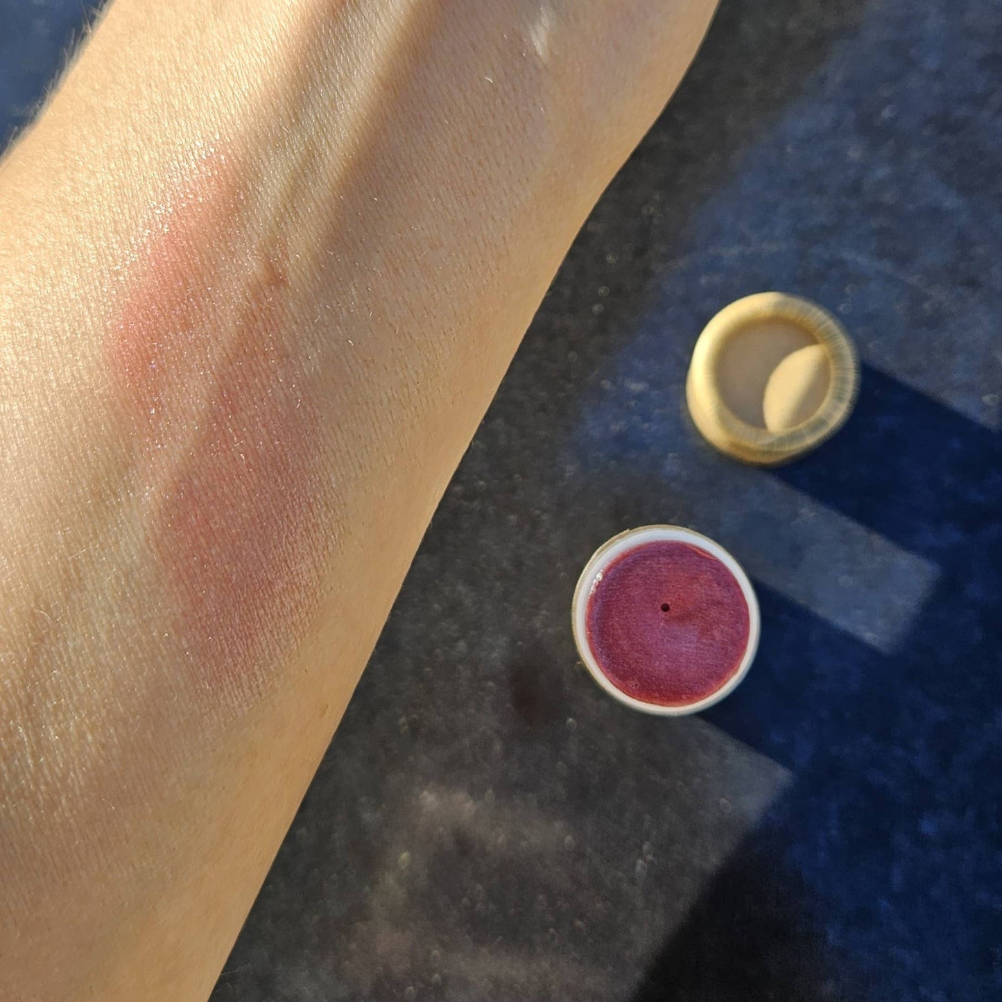 Close-up of a hand with pink blush and a small container of pink makeup on a dark surface.