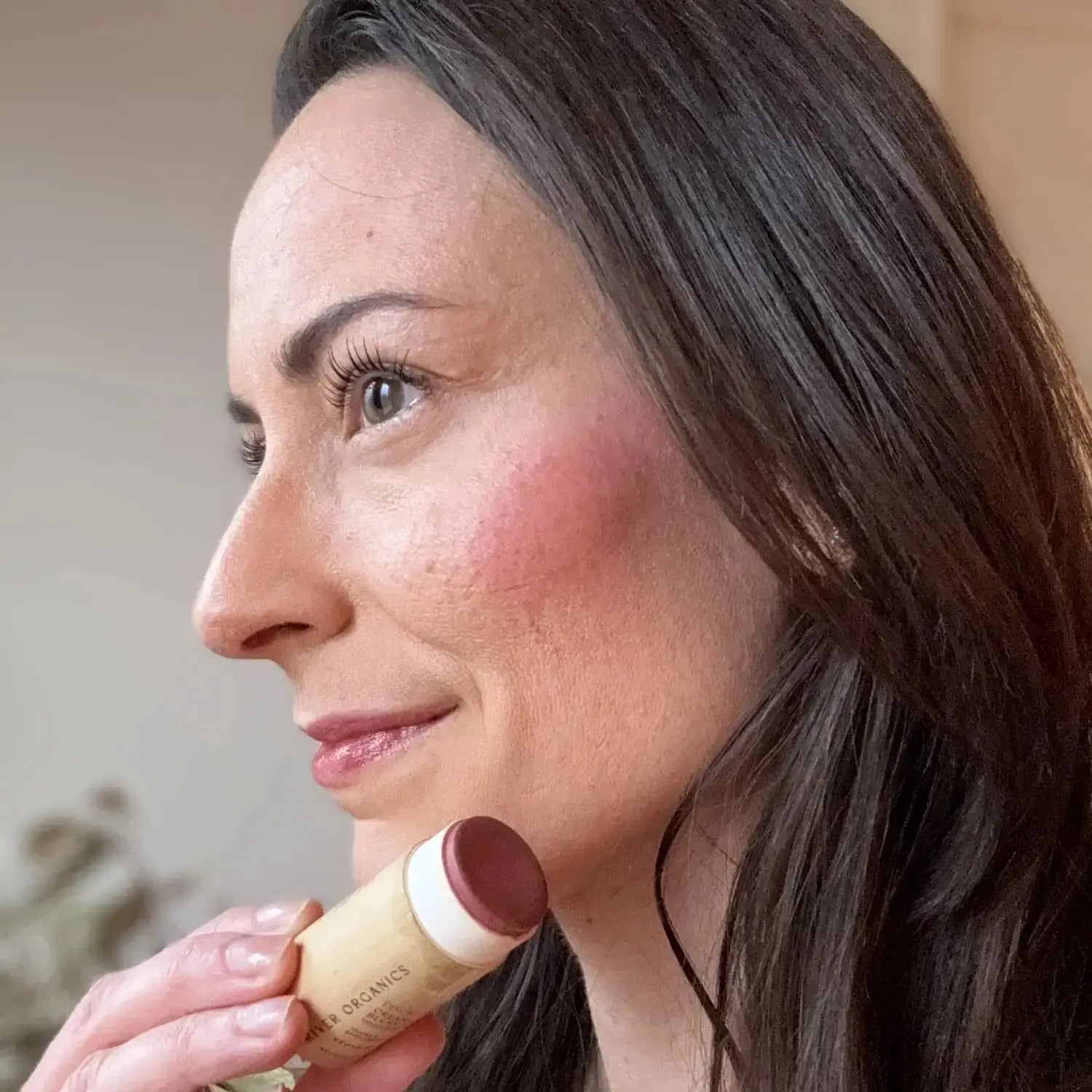 Woman applying Pinch Pink Organic Blush Stick to cheek, demonstrating smooth application and buildable color. Vegan and hydrating cream blush.