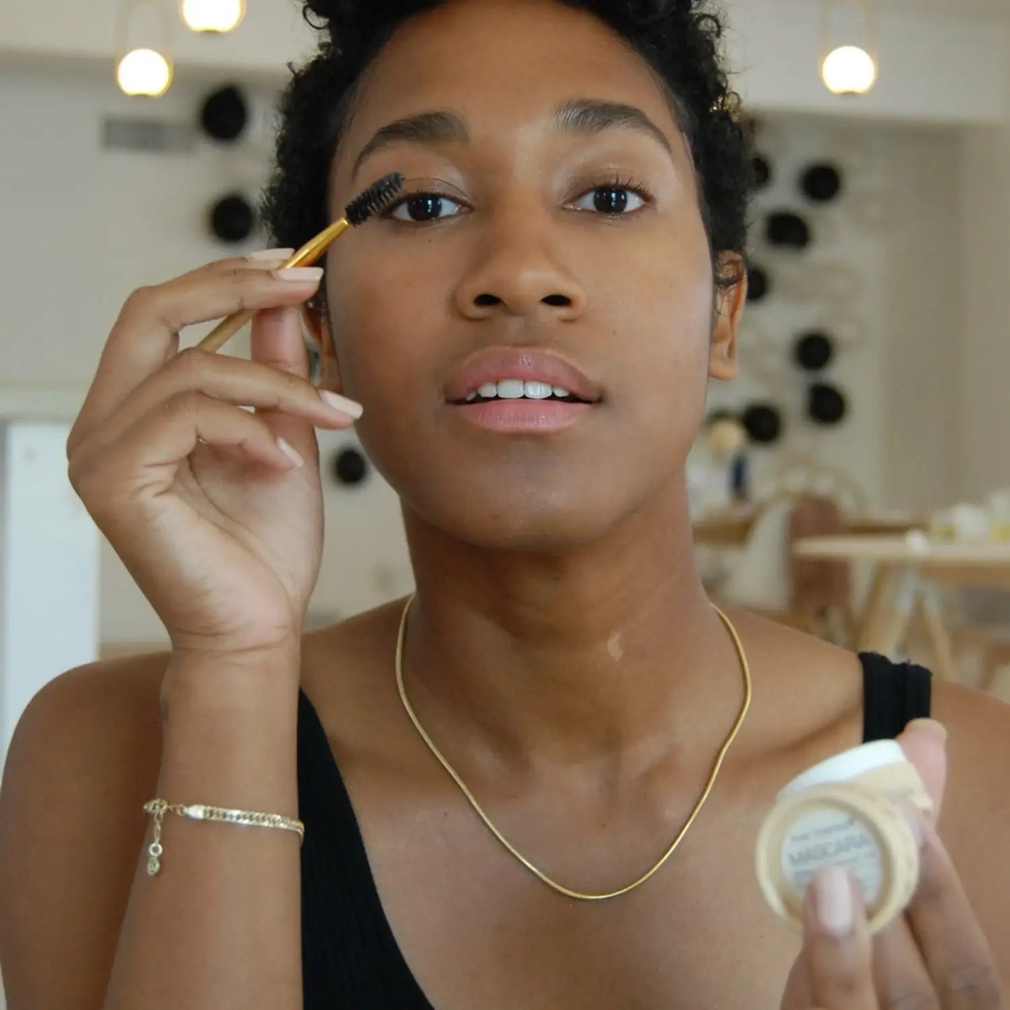 Woman applying cake mascara with a brush holding a cake mascara pot in a casual setting