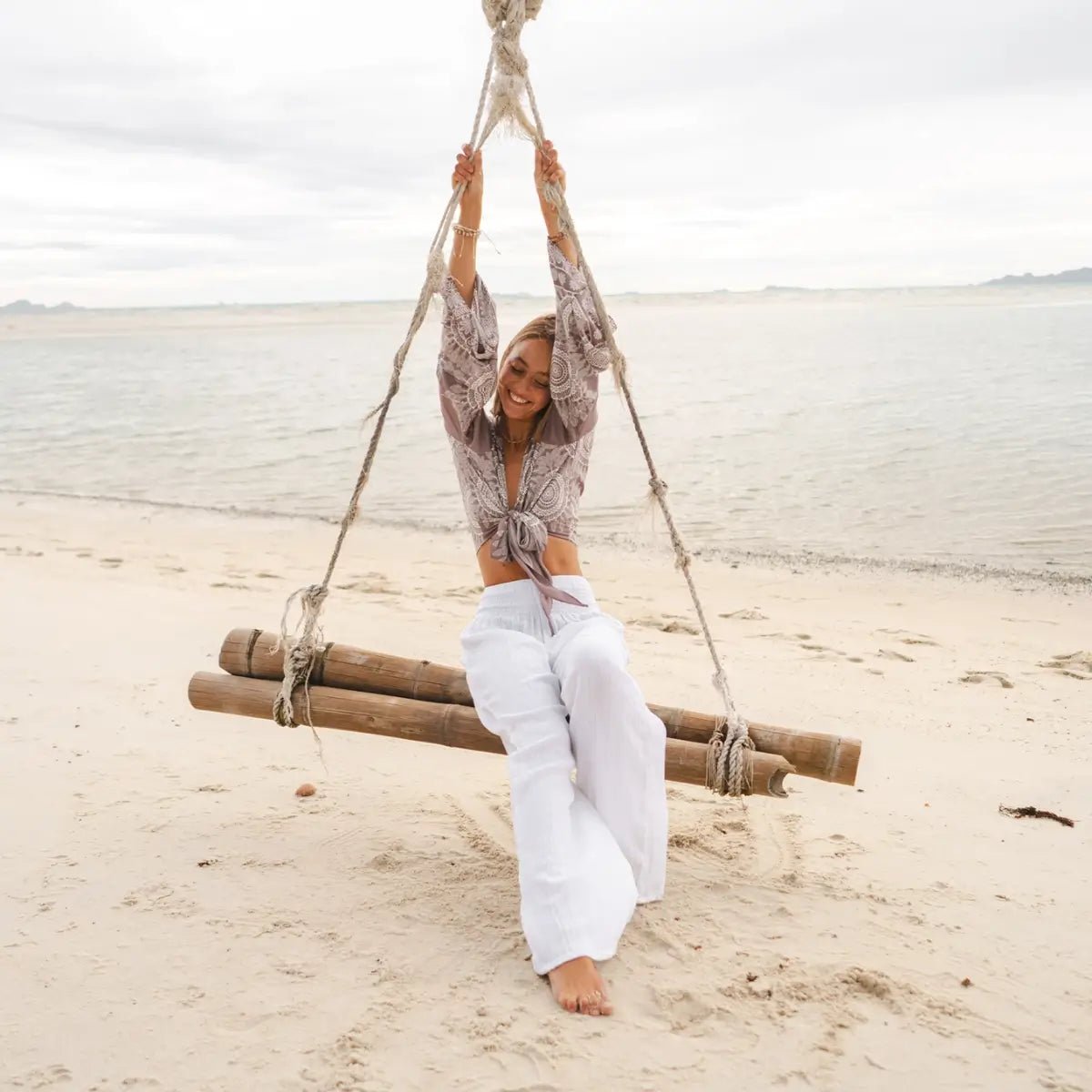 woman on a swing in the sand wearing lotus and luna white cotton beach pants - elle and willow boho beachwear collection