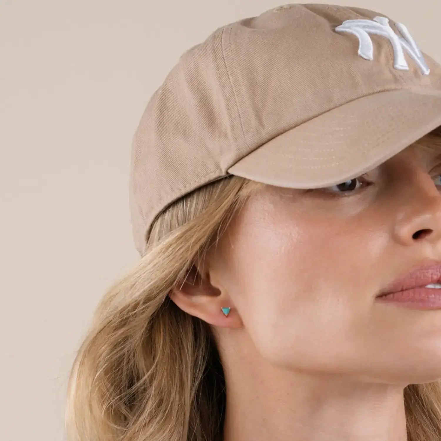 Close-up of a person wearing blue turquoise and yellow gold stud earrings with a beige NY Yankees cap