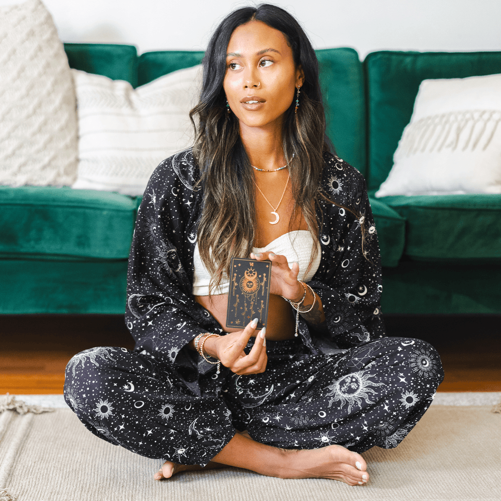 Woman in a black patterned outfit sitting on the floor holding a tarot card.