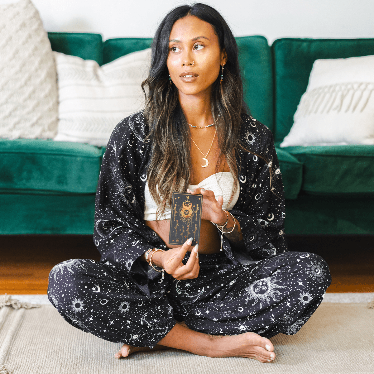 Woman in a black patterned outfit sitting on the floor holding a tarot card.