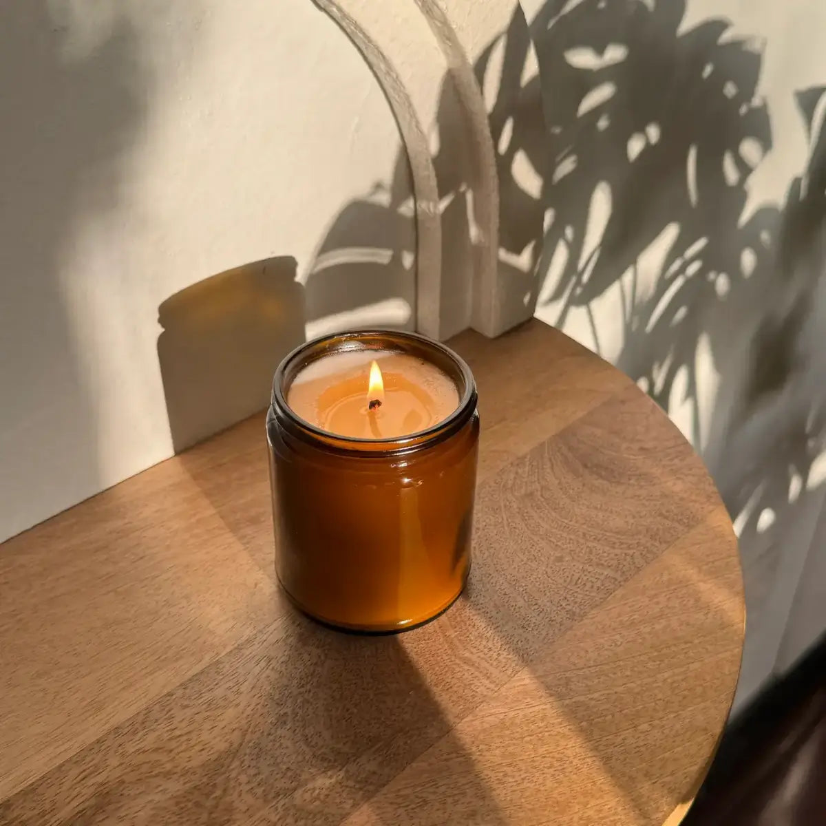 Amber glass candle with a lit wick on a wooden surface, casting shadows.