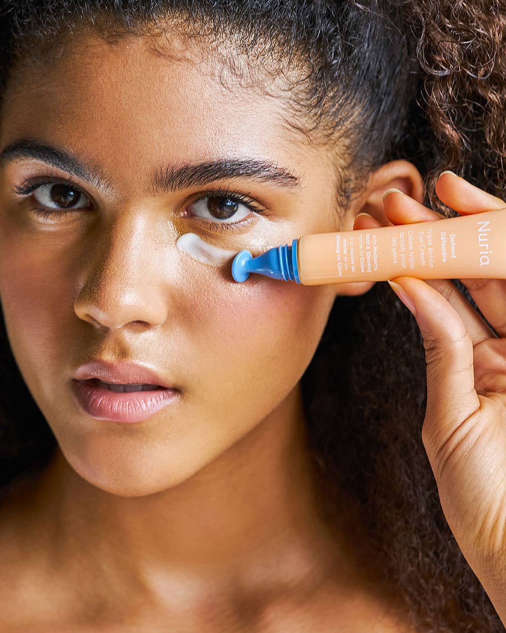 Model applying Nuria Eye Cream under her eye using the applicator, showcasing its brightening and puffiness-reducing benefits.