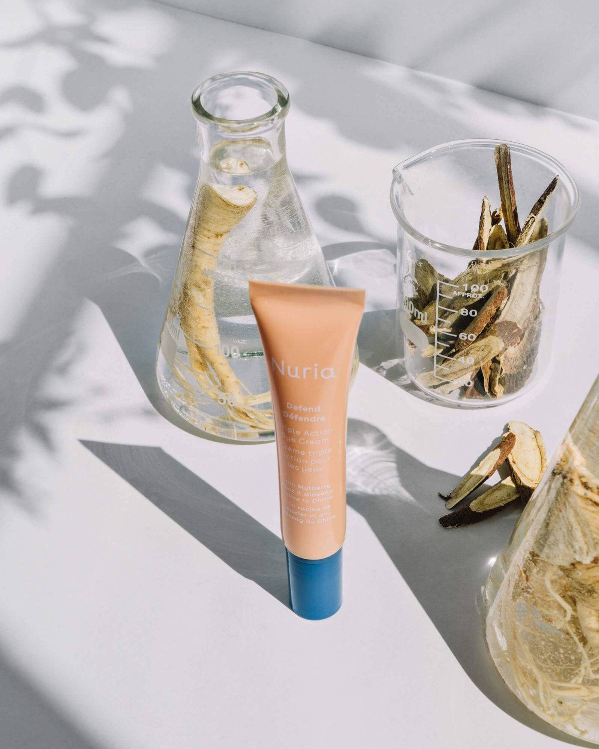 Nuria Eye Cream tube placed next to beakers with natural extracts, highlighting ingredients like Mulberry Root and Ginseng.