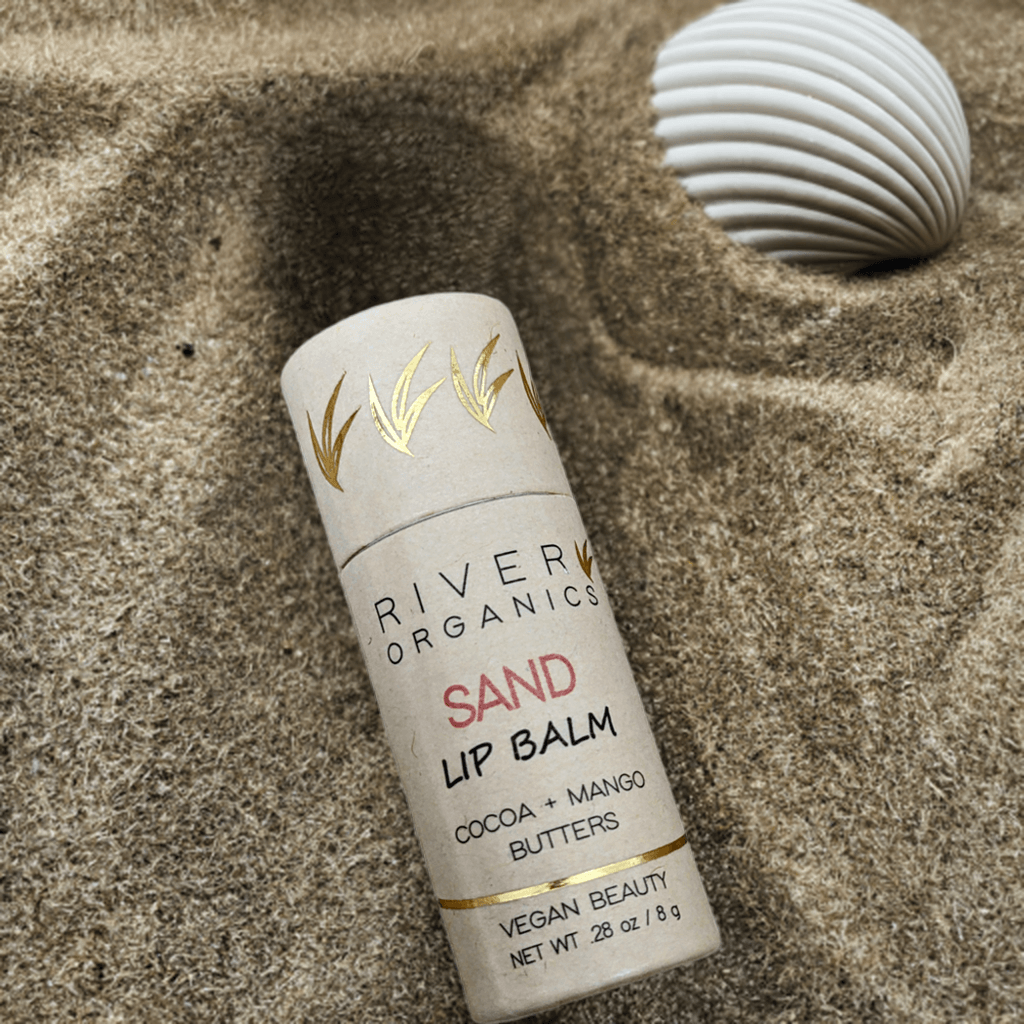 shimmering sand colored tinted lip balm in a zero-waste paper tube lying in the sand at the beach next to a seashell