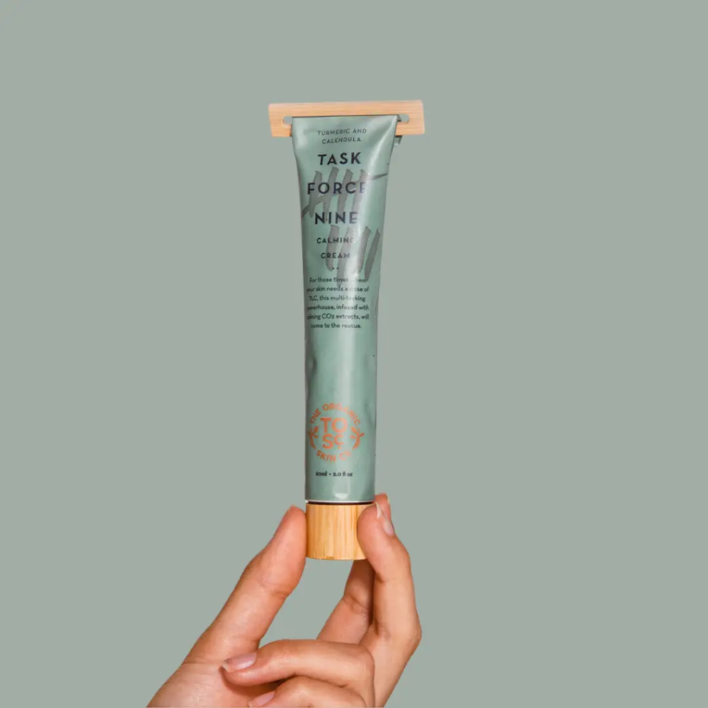 A hand holding a tube of The Organic Skin Co. Task Force Nine Aloe Calming Cream