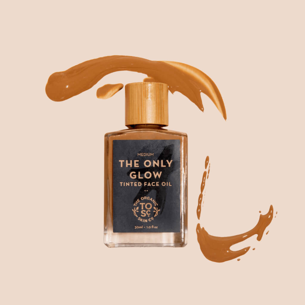 The Only Glow - Hydrating Organic Foundation Serum in a glass bottle with a wooden cap, surrounded by swirls of tinted face oil on a beige background.