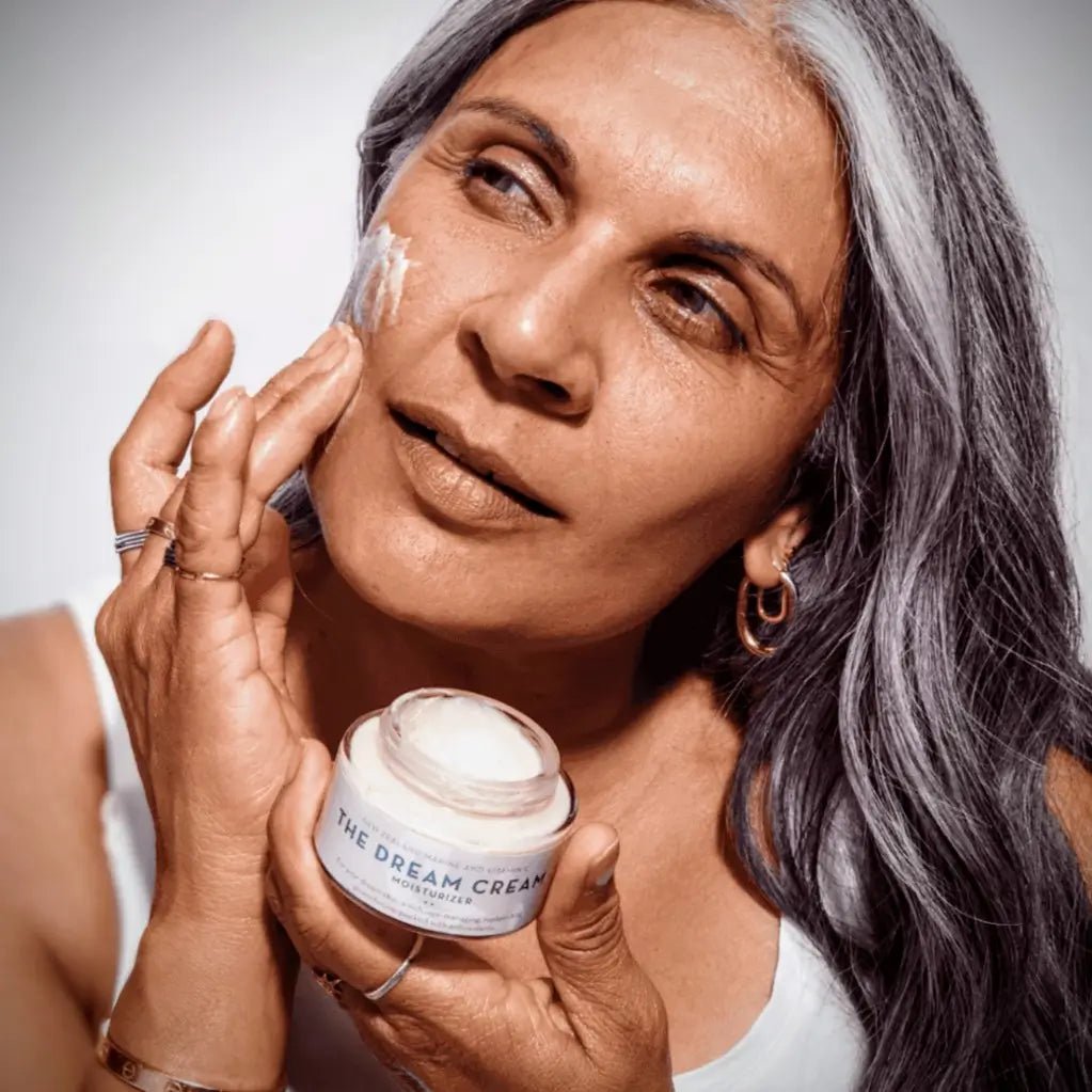Woman applying cream to her face with a jar labeled 'The Dream Cream' against a neutral background