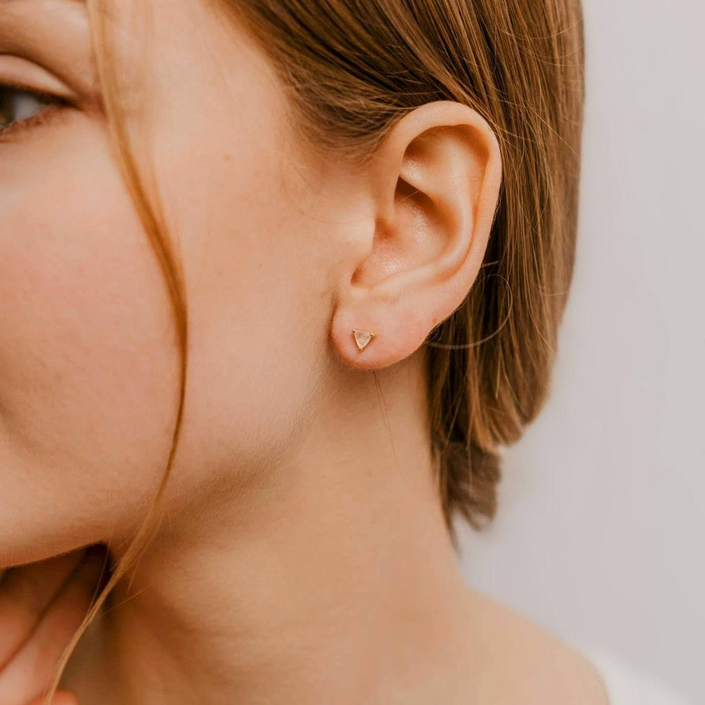 Mini Energy Gemstone Stud Earrings in 18k Yellow Gold featuring a triangular cut Rose Quartz gemstone worn on a woman's ear. Elegant and minimalist jewelry for everyday wear.