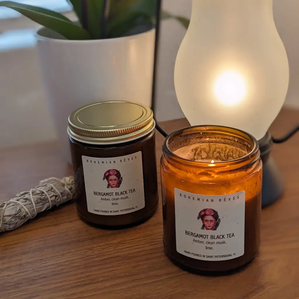 Two candles labeled 'Bergamot Black Tea' on a wooden surface with a lamp and plant in the background.