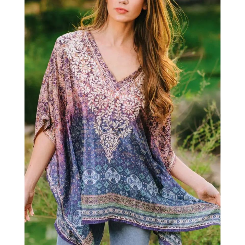 A woman wearing a loose, V-neck bohemian-style tunic top with a purple, blue, and white floral pattern, paired with blue jeans.