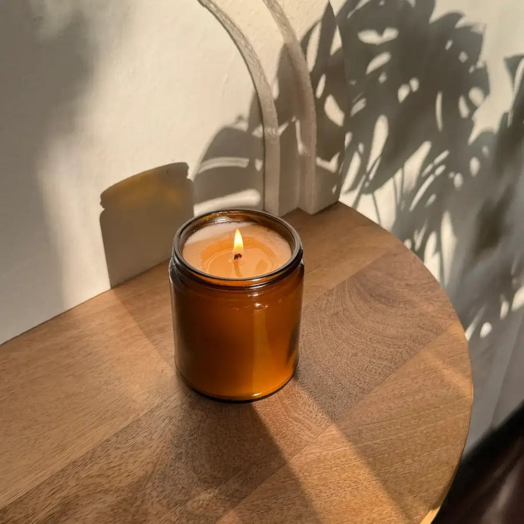 Bergamot black tea soy candle by Bohemian Reves in an amber glass jar with a lit wick on a wooden surface, casting shadows.