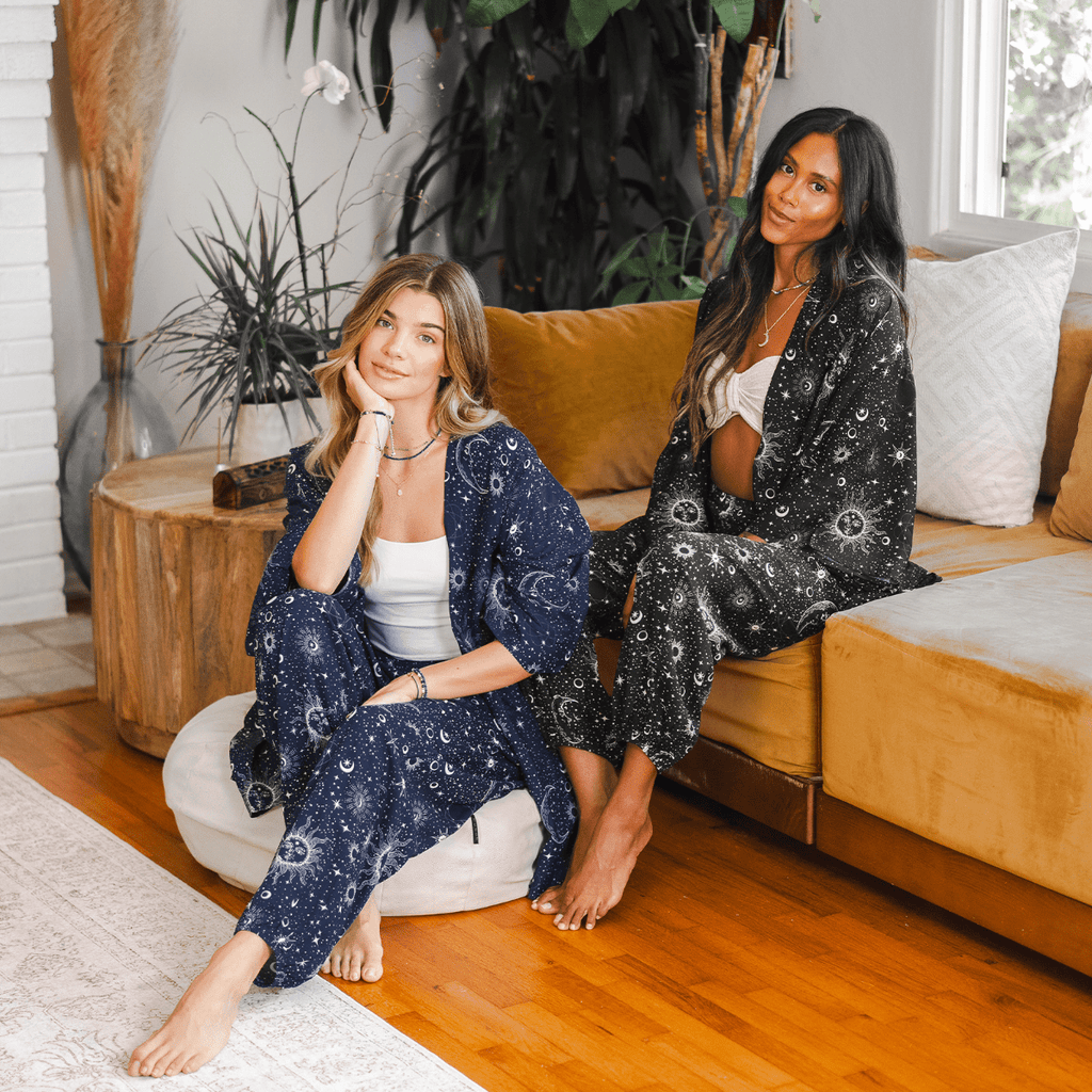 Two women in matching lotus and Luna cosmic Sky Kimonos and pants sitting on a couch in a cozy living room.