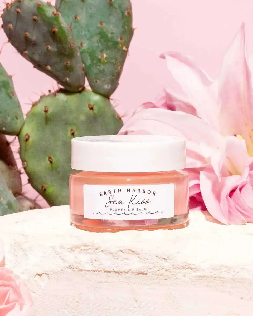 Earth Harbor SEA KISS lip balm jar displayed with cactus and pink flowers, highlighting its natural, organic ingredients for soft, plump lips.