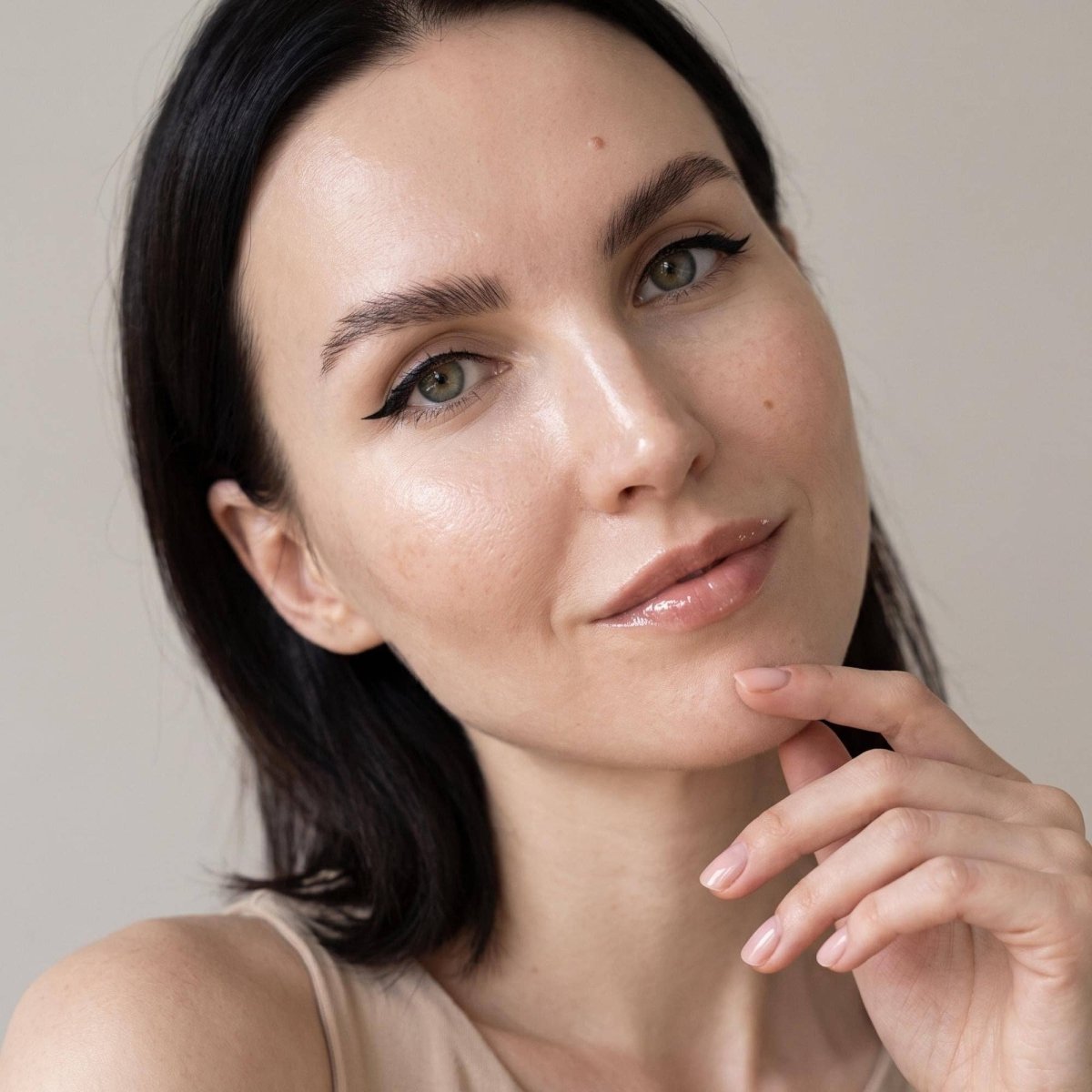 A close-up portrait of a woman with dark hair, wearing a beige sleeveless top against a soft beige background.