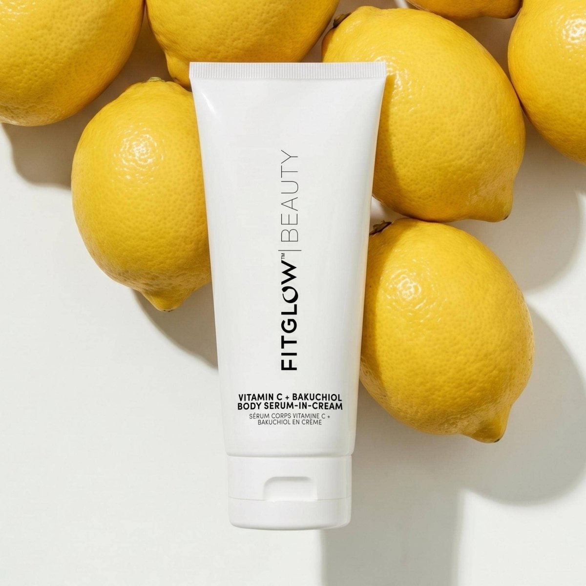 A white tube of FitGlow Beauty Vitamin C + Bakuchiol Body Serum-In-Cream surrounded by several bright yellow lemons.
