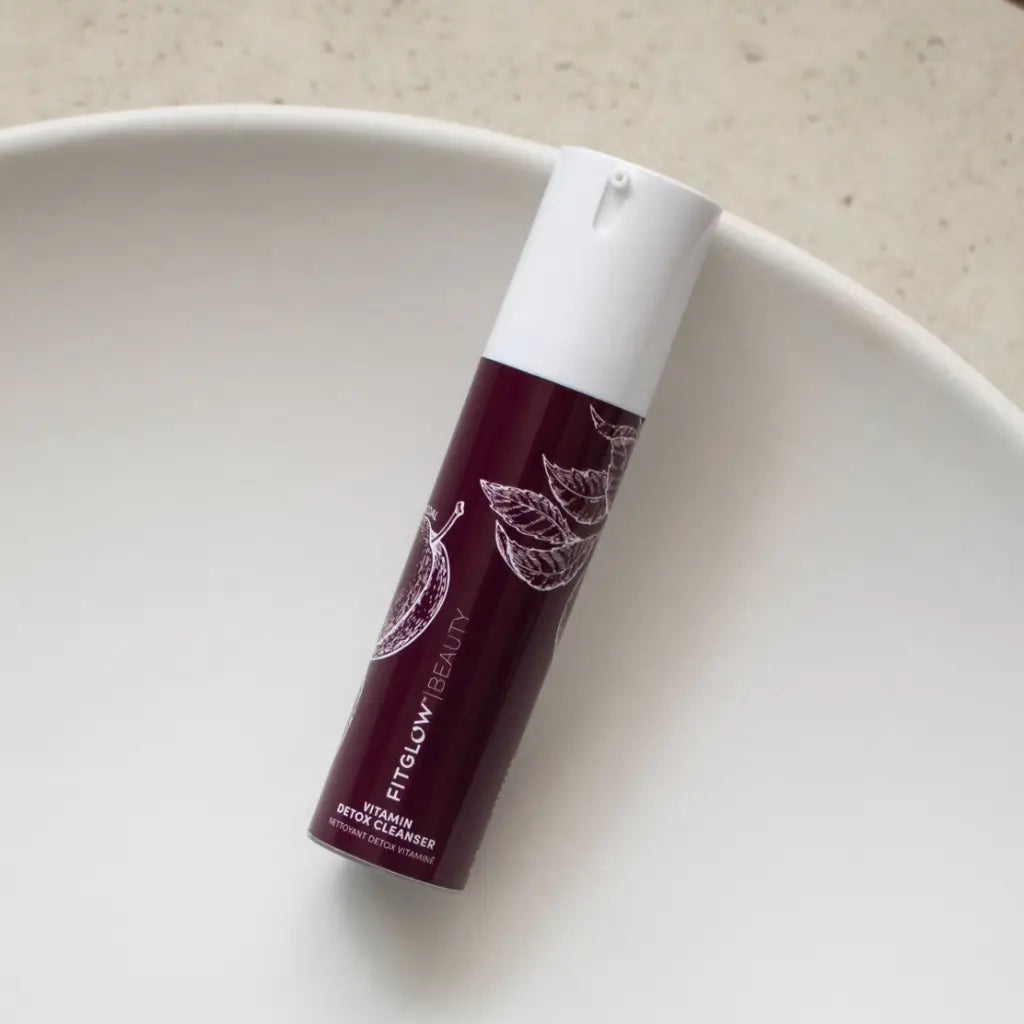 A bottle of FITGLOW BEAUTY Vitamin Serum Cleanser with a deep burgundy body and white cap, resting on a white ceramic dish against a light beige background.