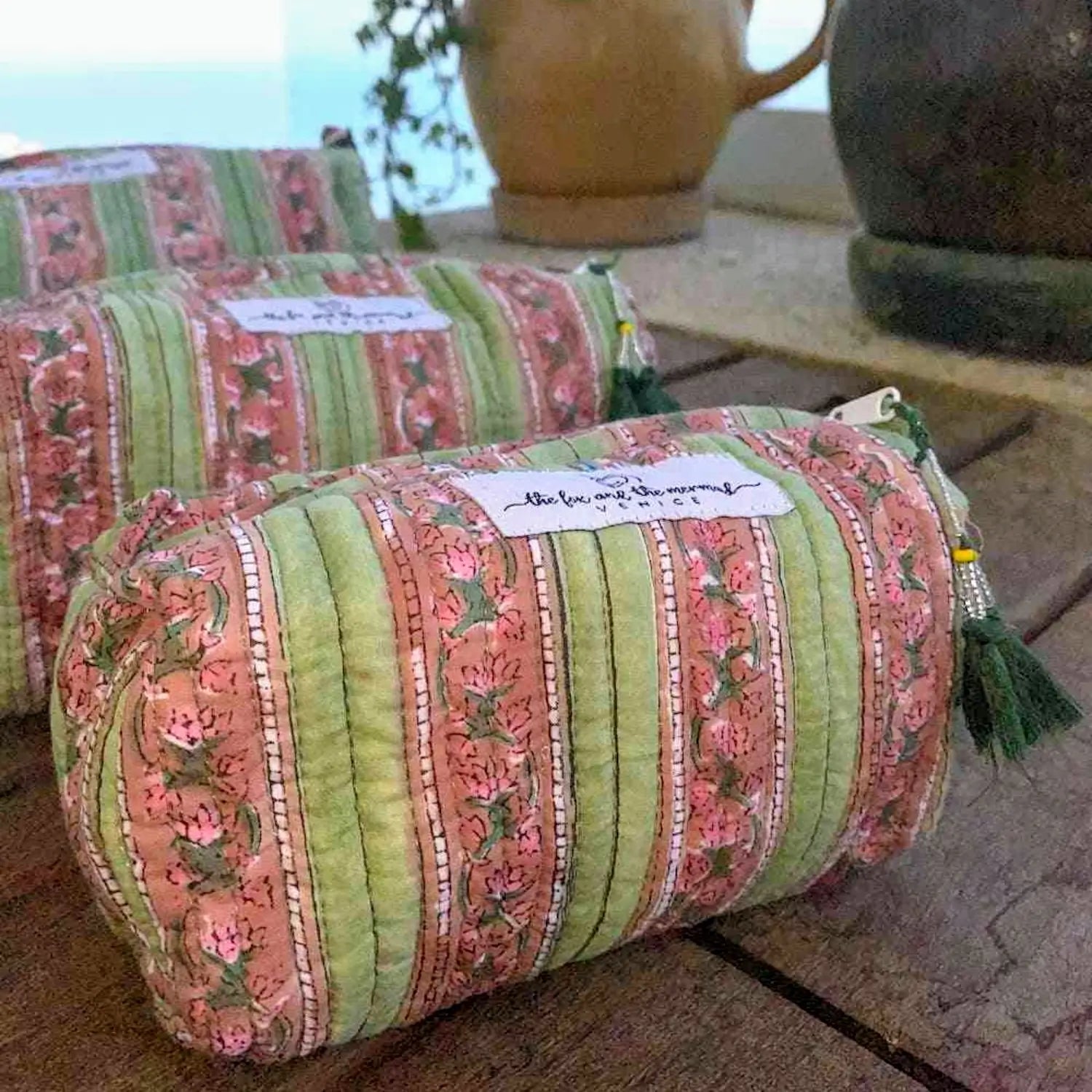 three fabric toiletry and cosmetic bags with zippered top and decorative tassels with pink and green floral striped pattern on a wooden surface.