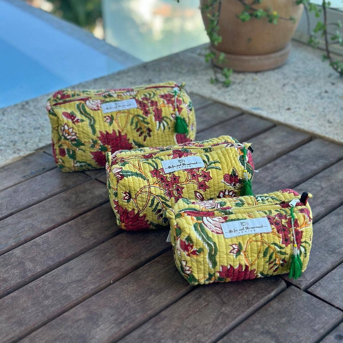 Three yellow floral-patterned cosmetic bags on a wooden surface with a blurred outdoor background.