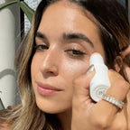 a woman applying a tube of light pink color correcting balm to her under eyes