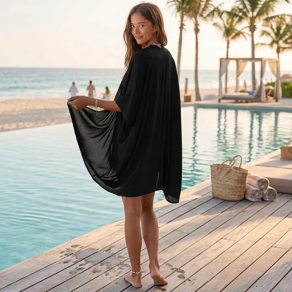 A woman wearing a flowing black kimono cover-up dress on a wooden deck beside a swimming pool.