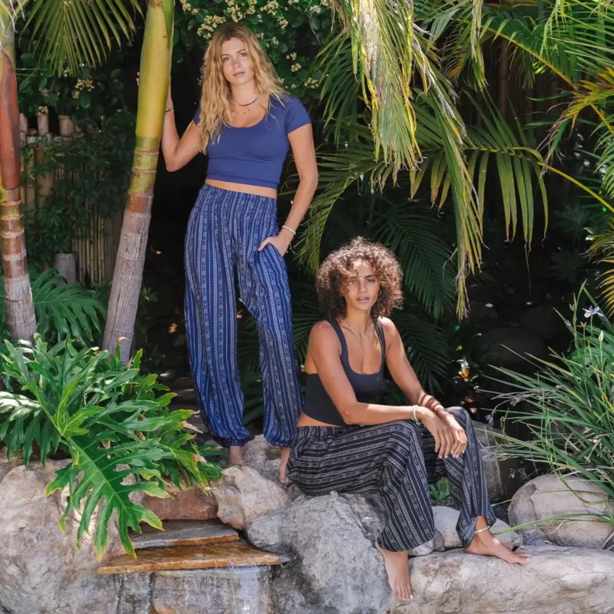 Two women wearing Lotus and Luna Boho Harem Pants, one in black striped and one in blue stripe in a tropical outdoor setting.