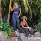 Two women wearing Lotus and Luna Boho Harem Pants, one in black striped and one in blue stripe in a tropical outdoor setting.