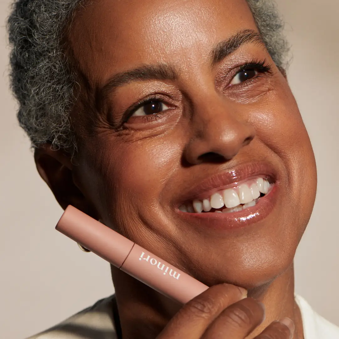 a woman with darker skin and dark eyes holding a tube of minori mother of lashes lengthening brown mascara with the defining bristle brush