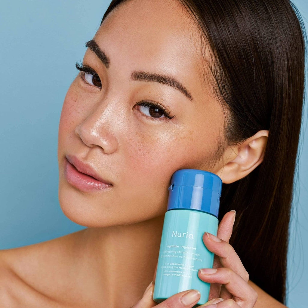 Woman holding a bottle 'Nuria Hydrate' micellar water against a blue backgroundof