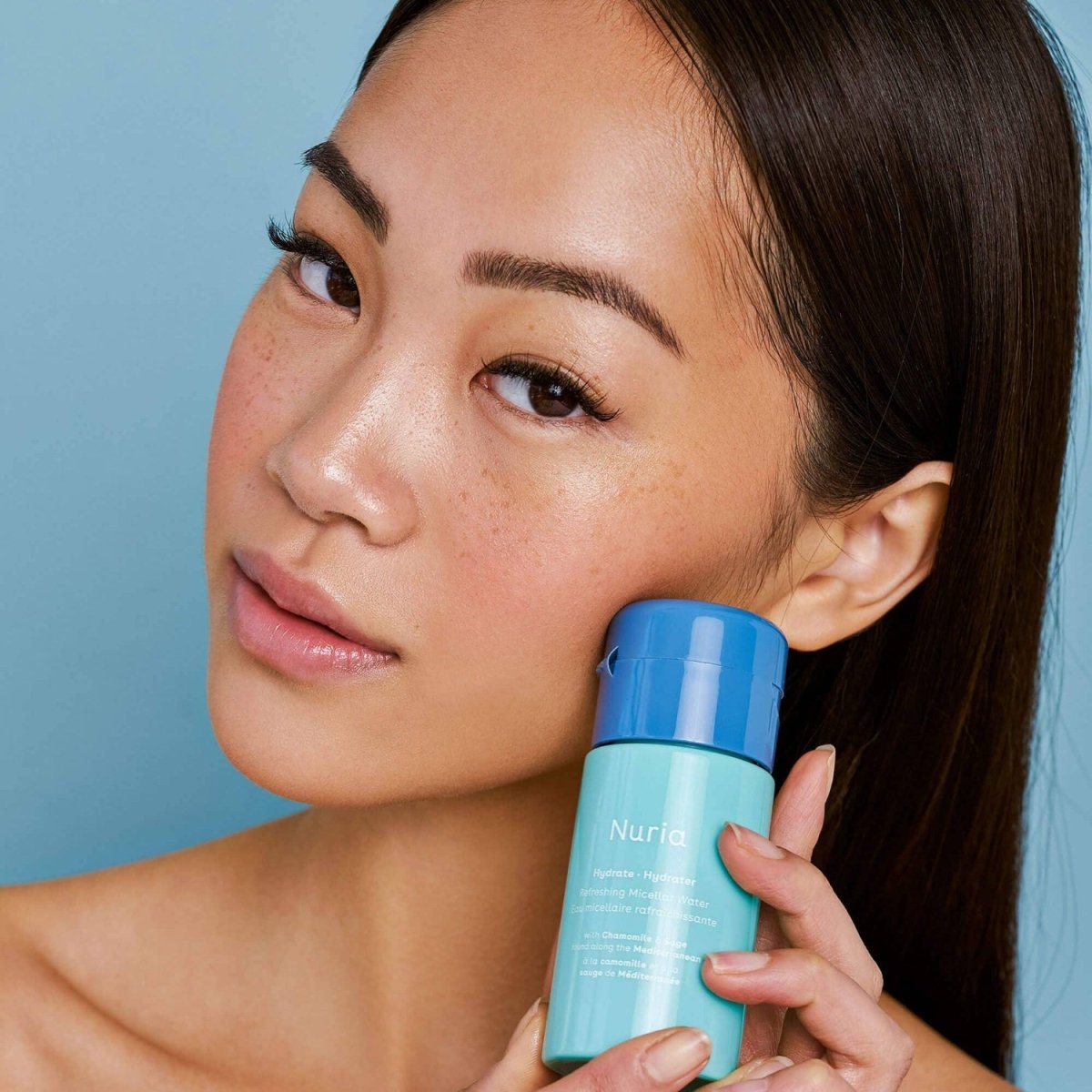 Woman holding a bottle 'Nuria Hydrate' micellar water against a blue backgroundof