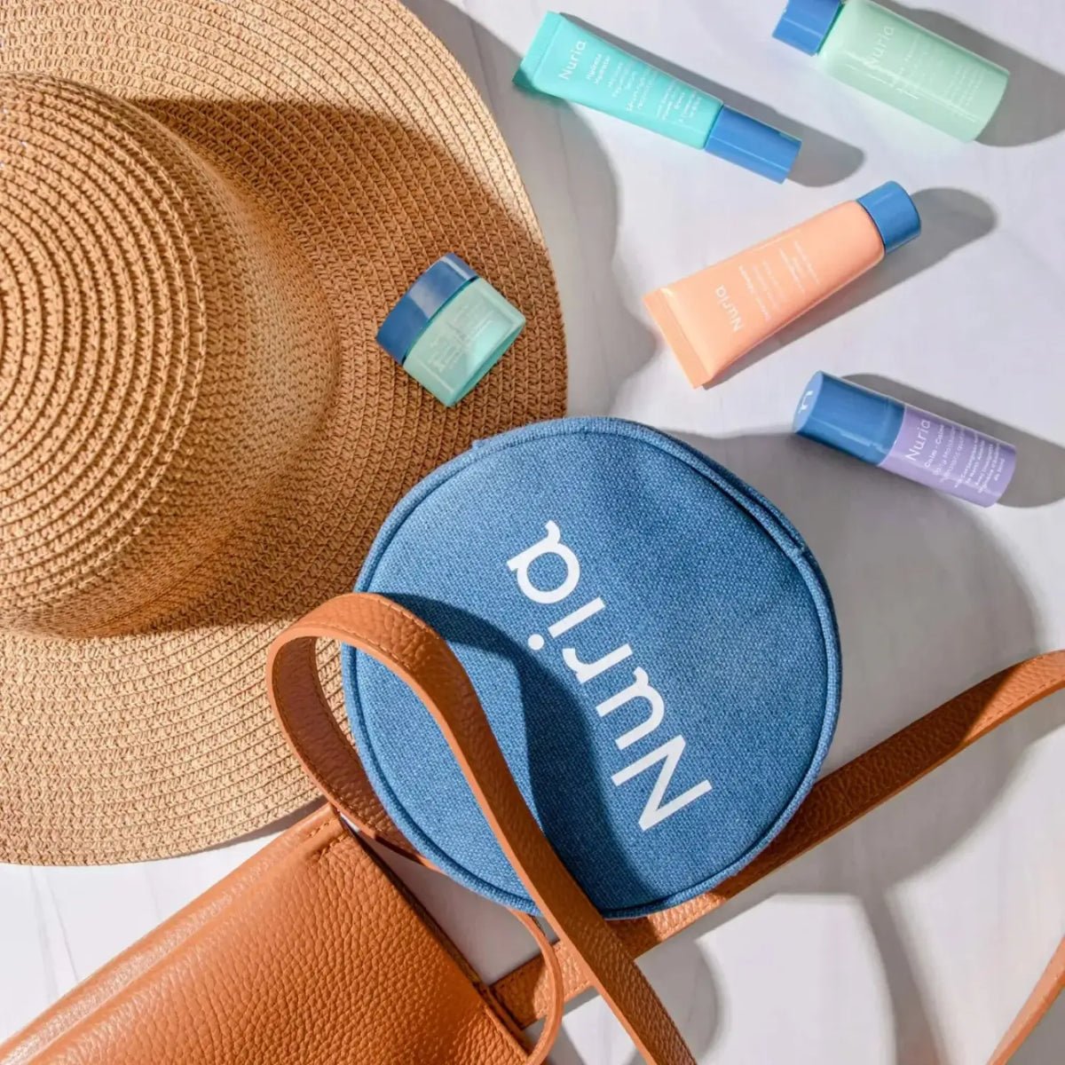 Blue toiletry pouch with 'Nuria' branding, straw hat, and skincare bottles on a white background