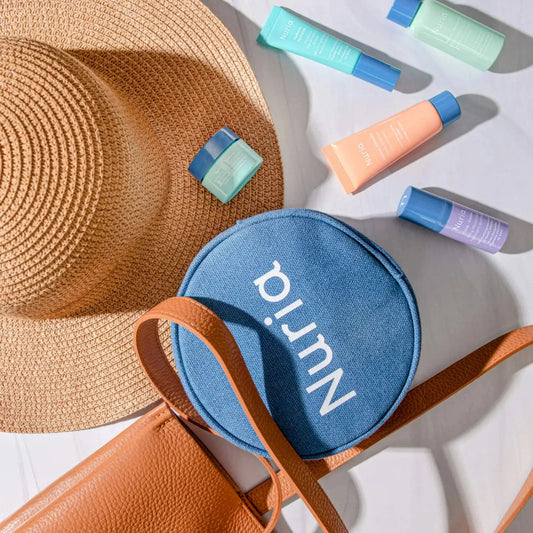 Blue circular toiletry bag with 'Nuria' branding, sun hat, and assorted travel size bottles of Nuria skincare products on a white background