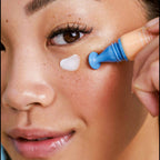 Close-up of a model applying Nuria Eye Cream under her eye, showcasing its targeted applicator and brightening benefits.