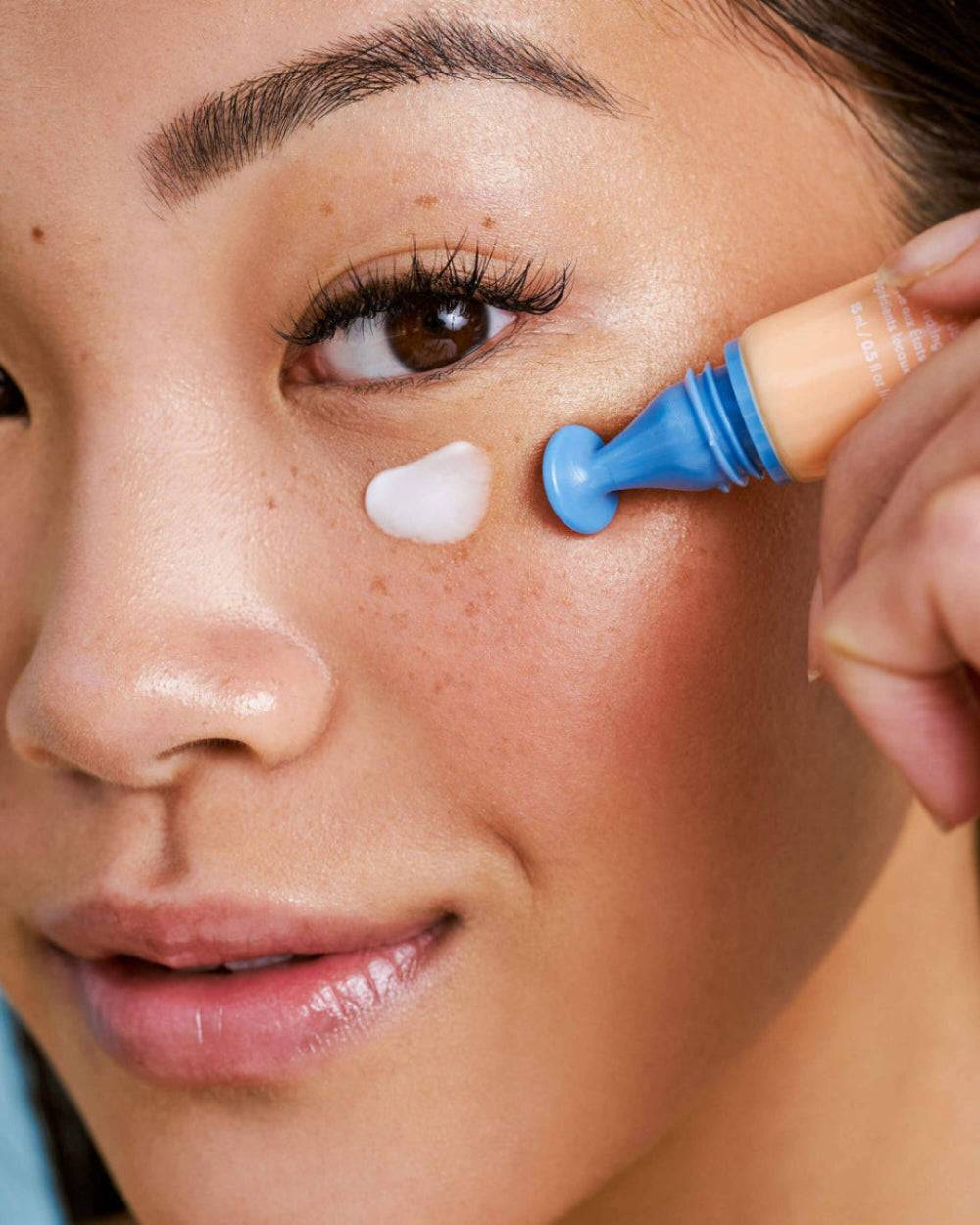 Close-up of a model applying Nuria Eye Cream under her eye, showcasing its targeted applicator and brightening benefits.