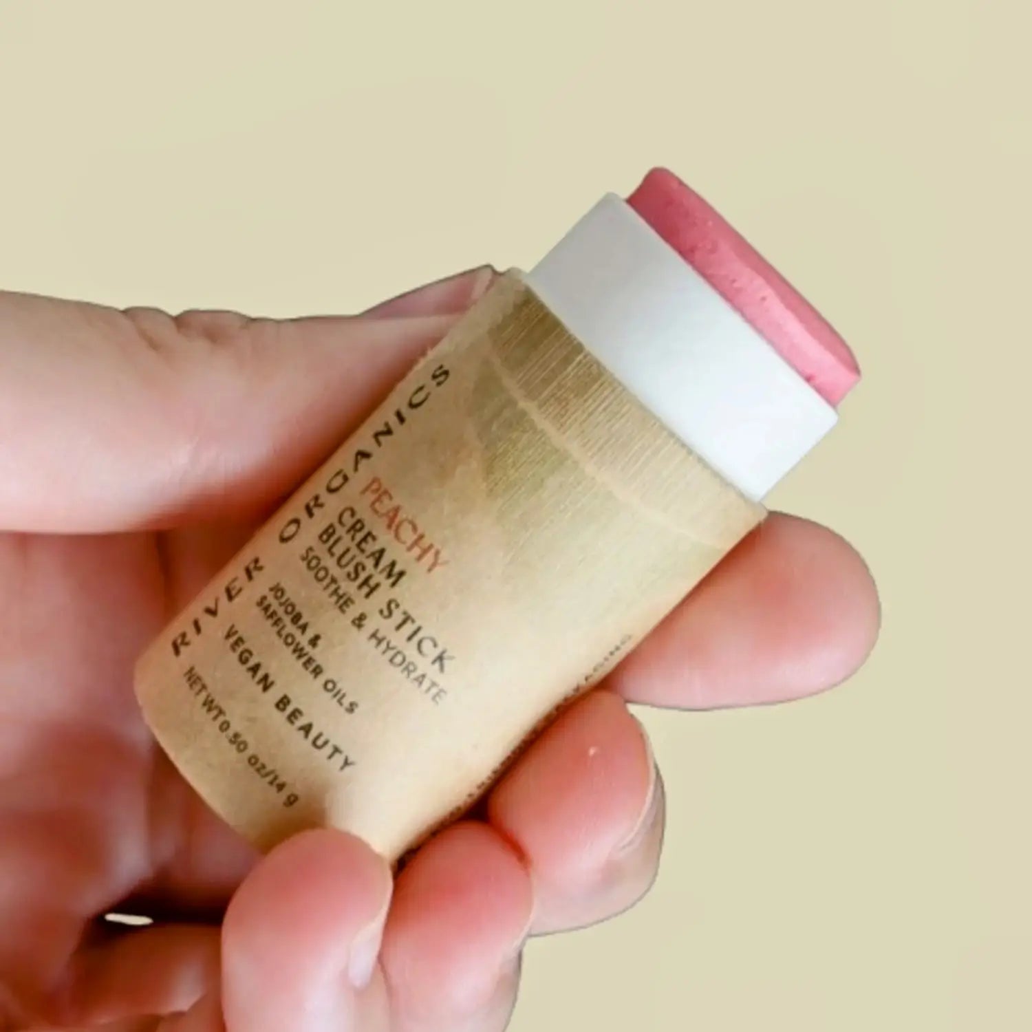 Hand holding a cream blush stick in a peach color with a beige background