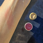 Close-up of a wrist with a swipe of Guava Pink lip stain  next to the pink lip balm tube on a dark surface.