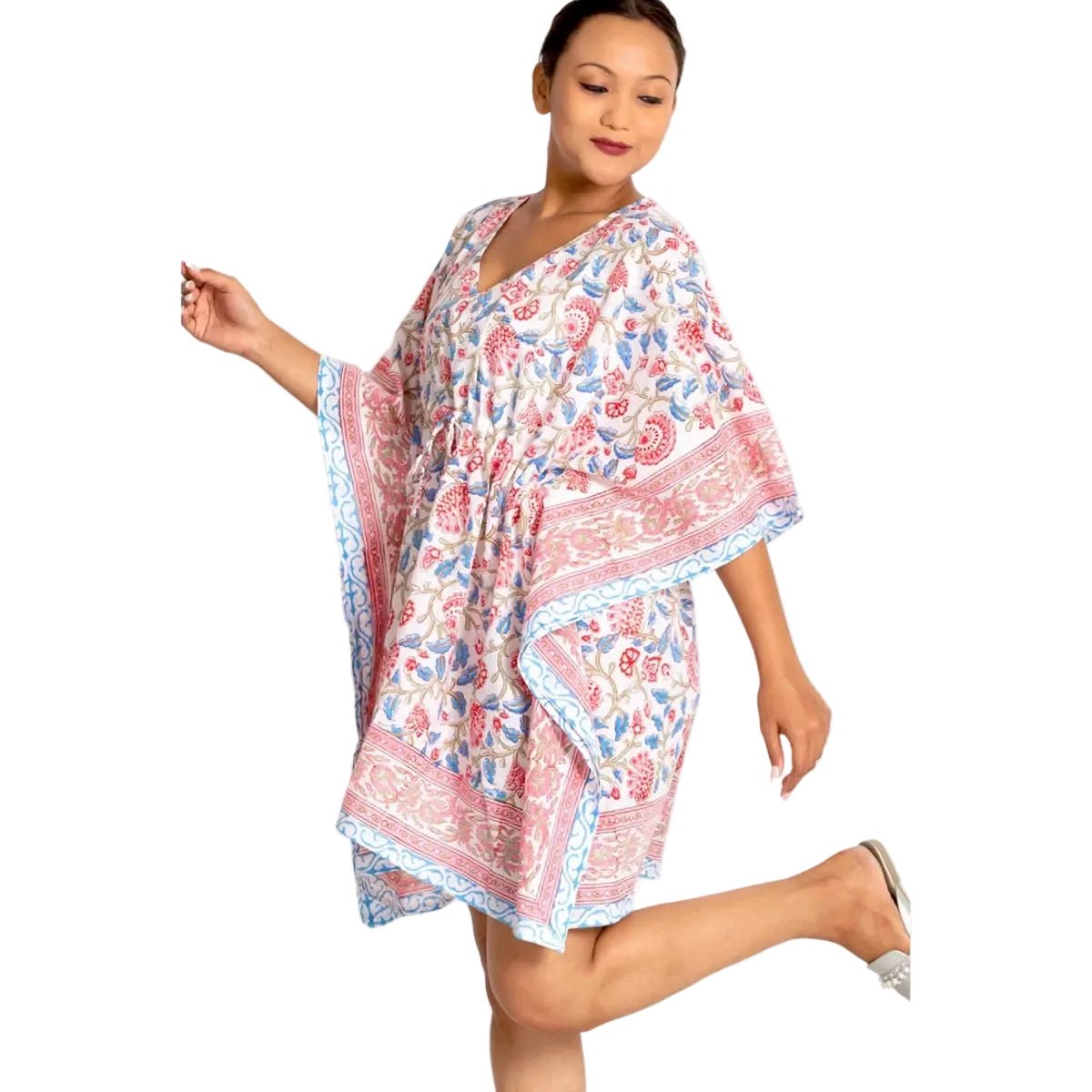 A woman modeling a short kaftan dress with a pink, blue, and beige floral and paisley print, set against a plain white background.