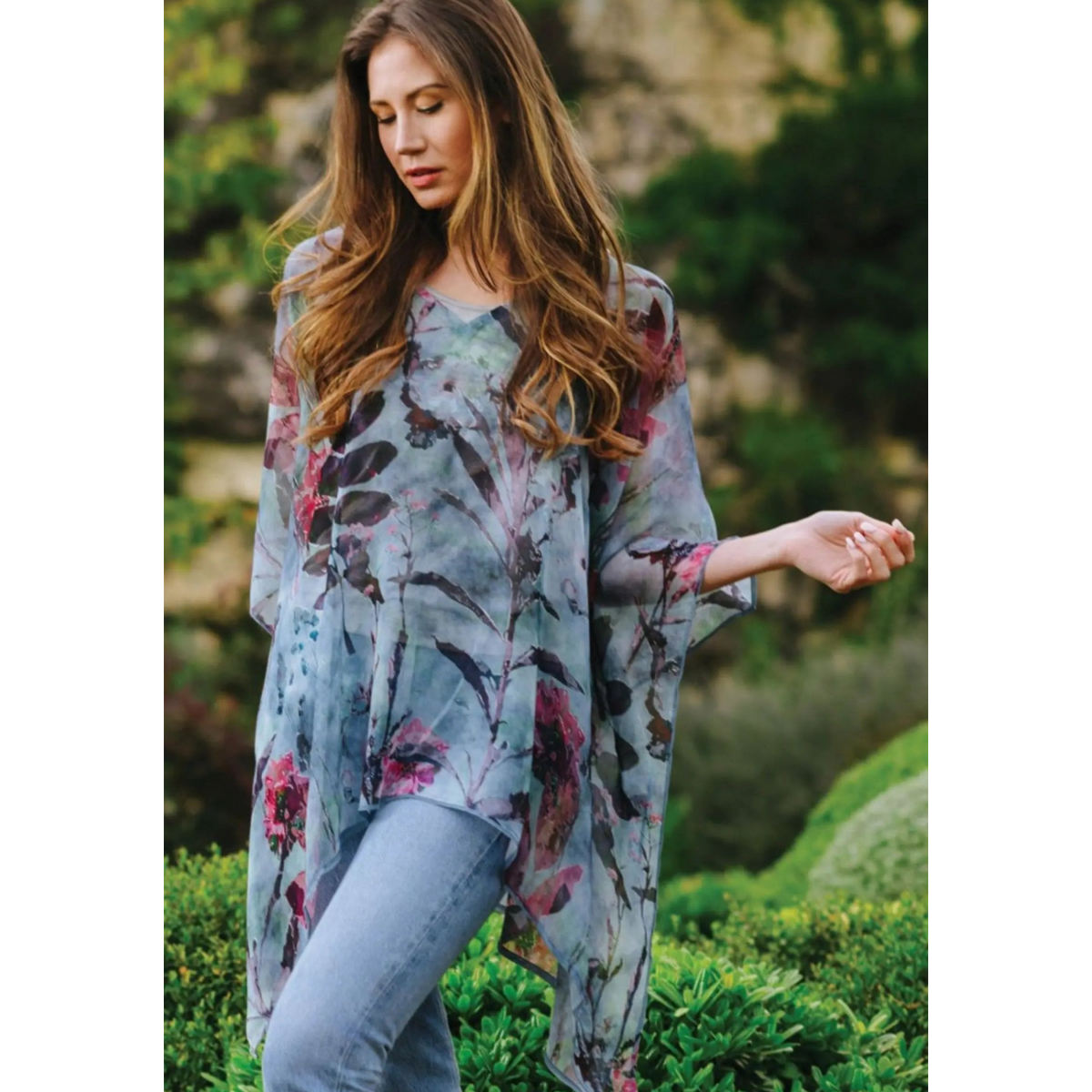 A woman wearing a loose, semi-sheer floral tunic top in muted blue-gray tones with pink and dark purple floral prints, paired with light blue jeans, standing outdoors in a garden setting.