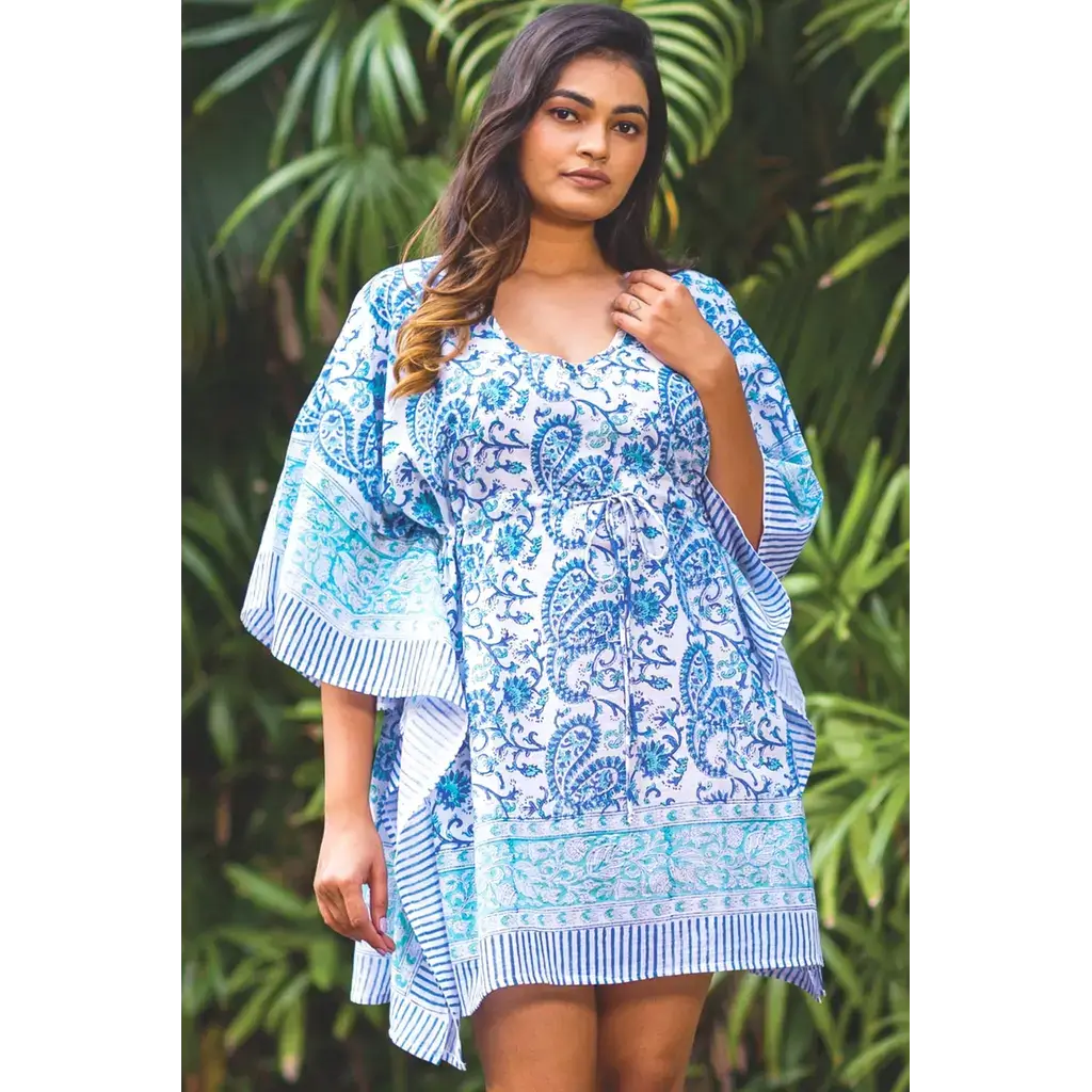 A woman wearing a blue and white paisley print kaftan dress with striped flutter sleeves, standing outdoors in front of a green tropical foliage background.