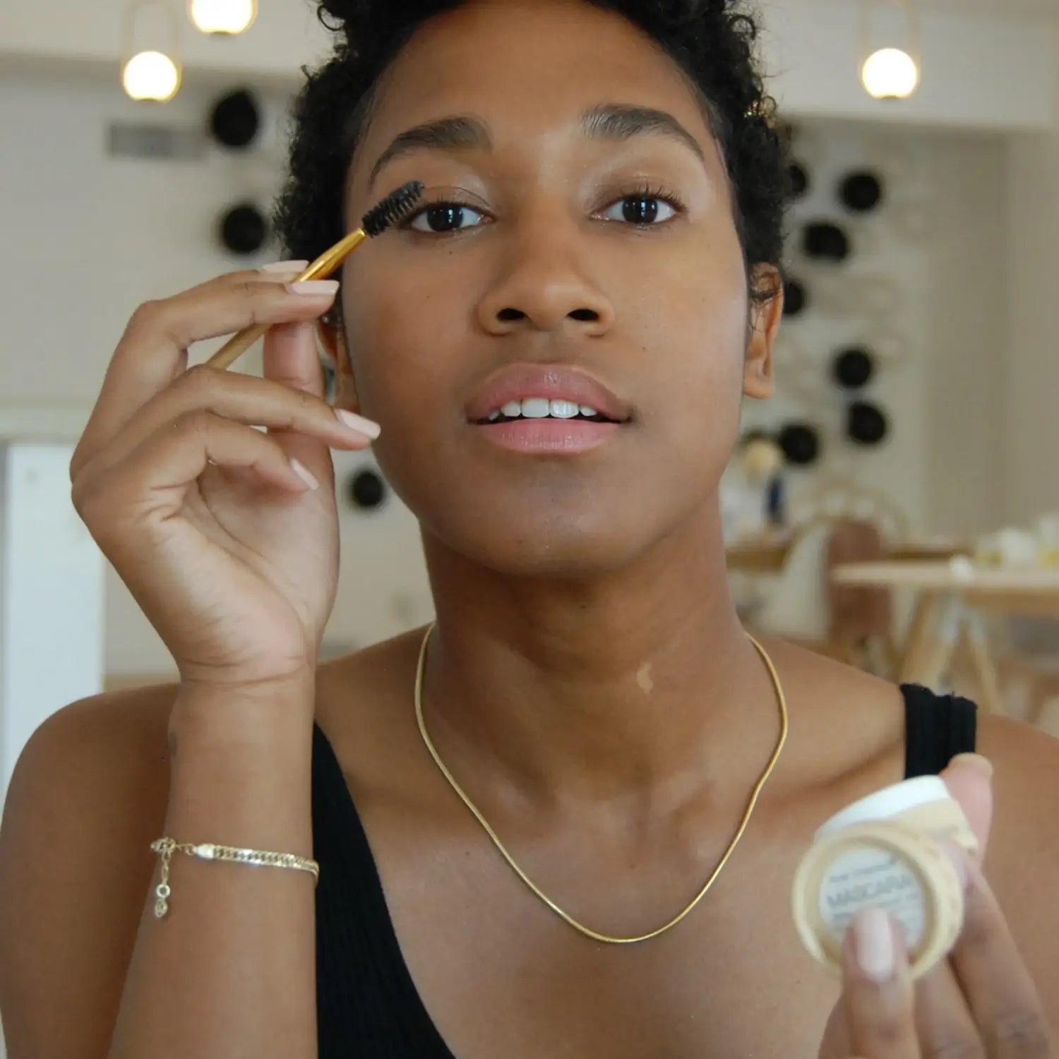 Woman applying cake mascara with a brush holding a cake mascara pot in a casual setting